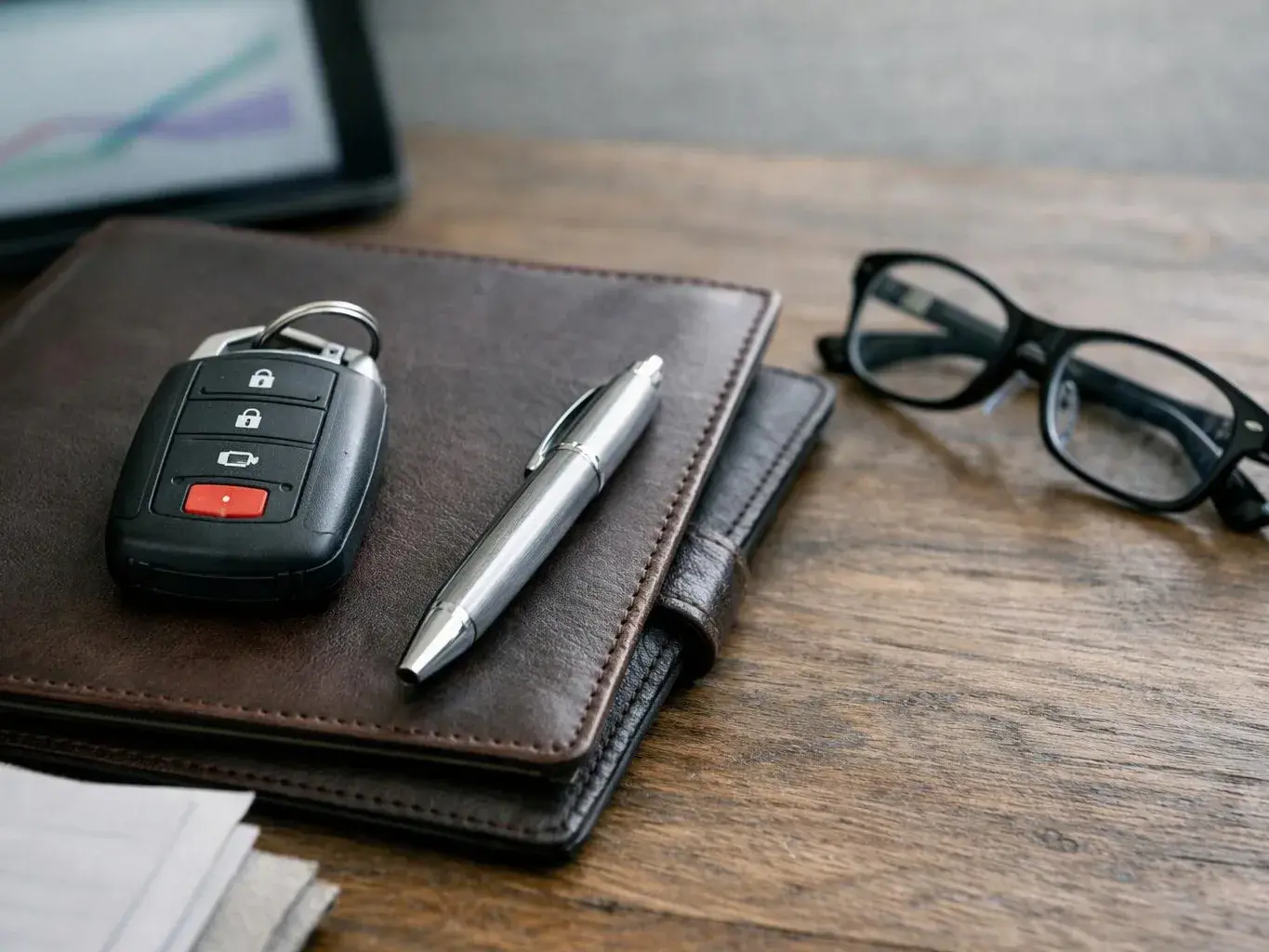 A car key fob, silver pen, and black glasses rest on stacked leather notebooks on a wooden desk