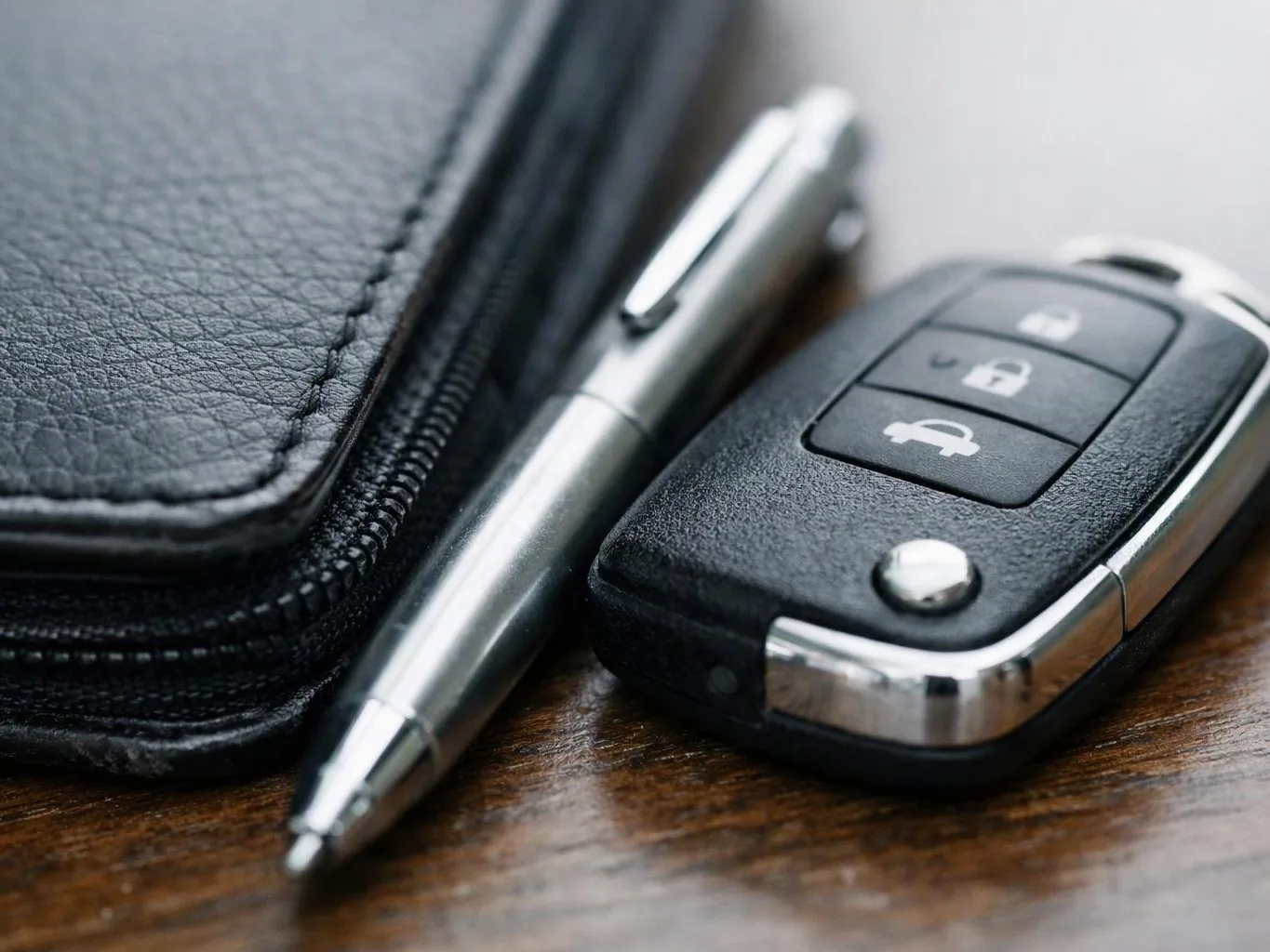 A car key fob, silver pen, and black zippered wallet rest on a wooden table