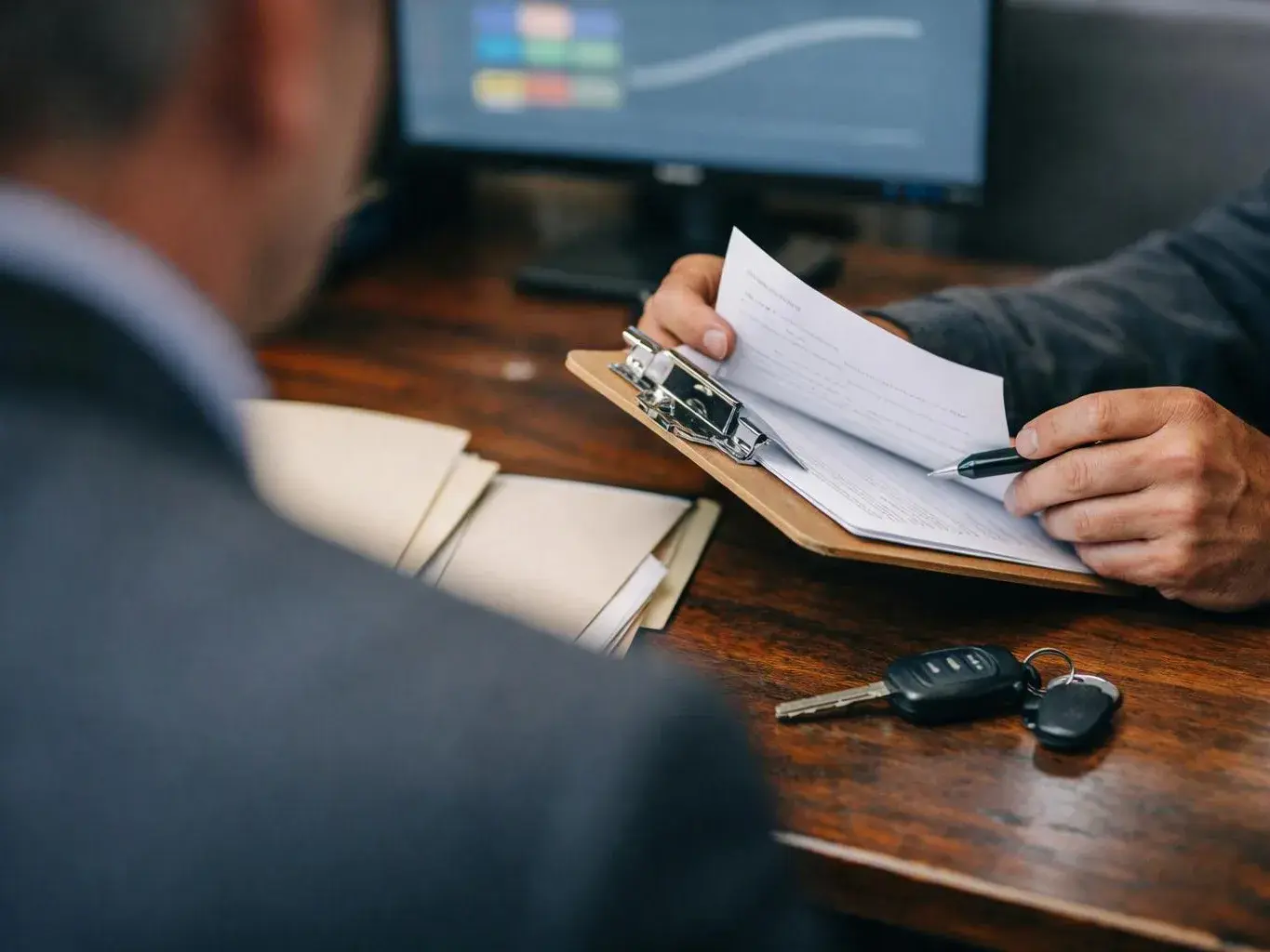 A person signs papers on a clipboard across a desk, with car keys nearby and a blurred monitor in the background