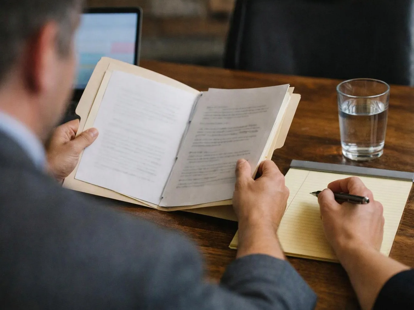 A person studies an open book and writes notes at a wooden table with a glass of water nearby