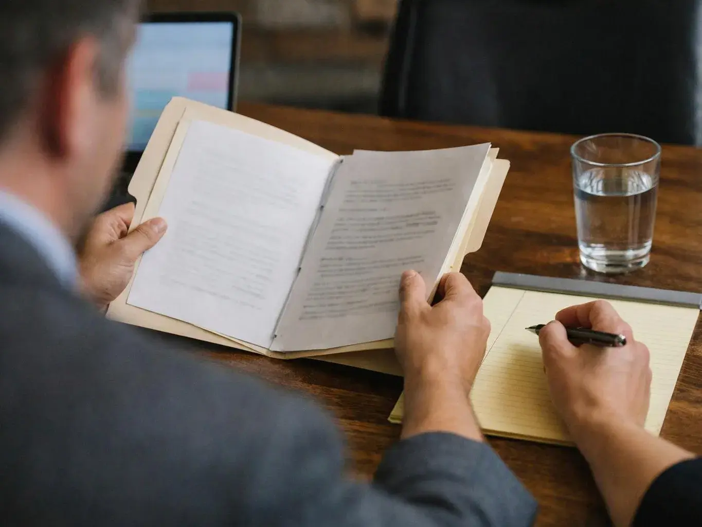 A person at a wooden table reads a paper document while another hand writes in a notepad beside a glass of water