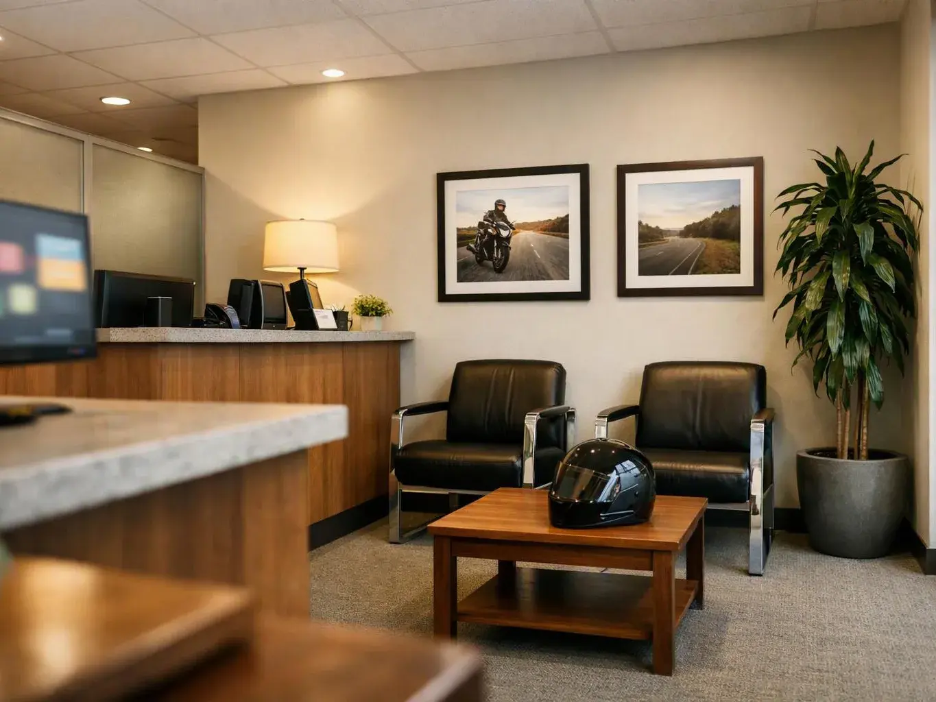 A quiet office waiting area has black chairs, a coffee table with a helmet, framed road photos, and a potted plant