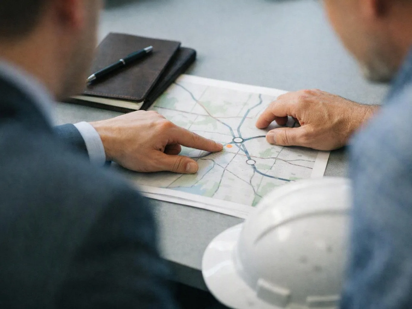 Two people point at a printed map on a table, with a notebook and pen nearby